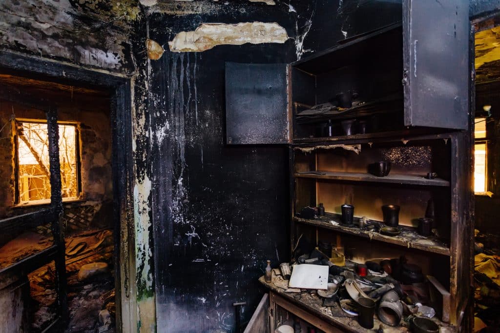 Fire-damaged house interior with charred walls, burned shelving, damaged furniture, and debris covering the floor.