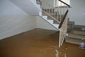 A home interior is severely flooded with muddy water covering the floor and surrounding a staircase.