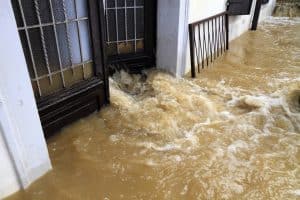 Muddy floodwater rushes into a building through the entrance, submerging the doorway and surrounding walkway.